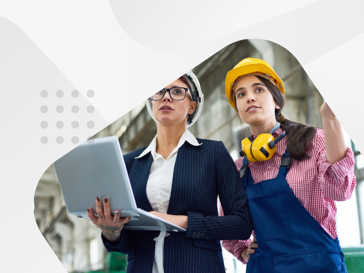 Two women wearing hard hats and holding laptops while working on a construction site.