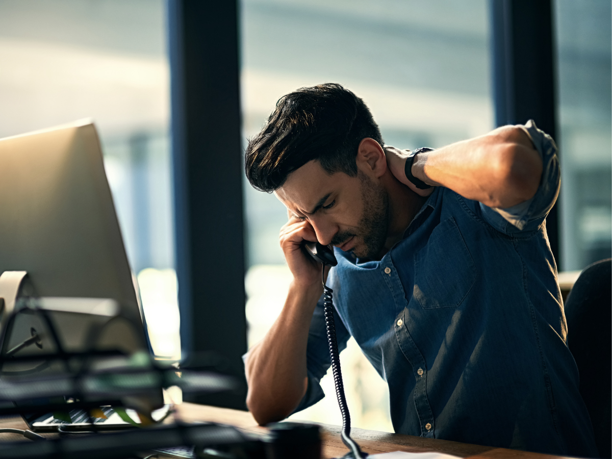 A man sitting at a desk with his head on the phone, looking stressed and overwhelmed.