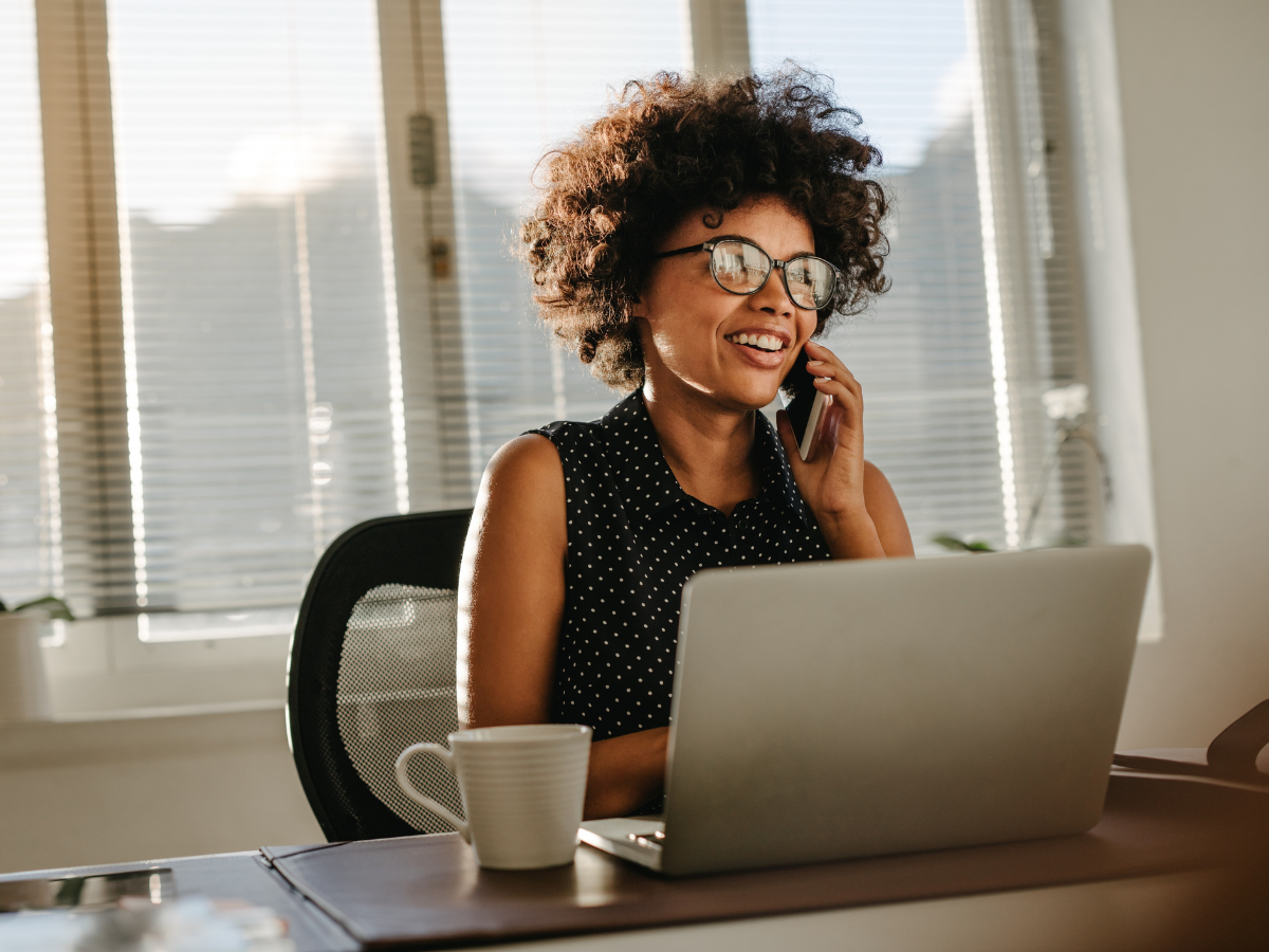 A woman in glasses and a polka dot shirt talking on the phone at her desk.