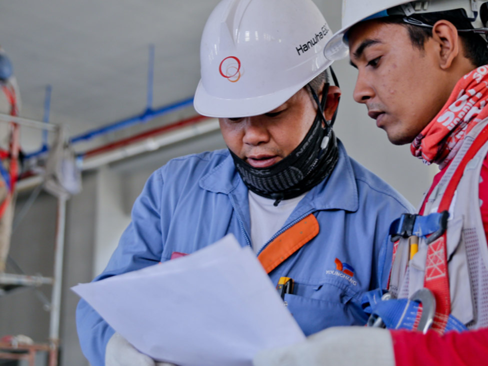 Two men indoors, wearing construction hard hats, looking at a piece of paper.