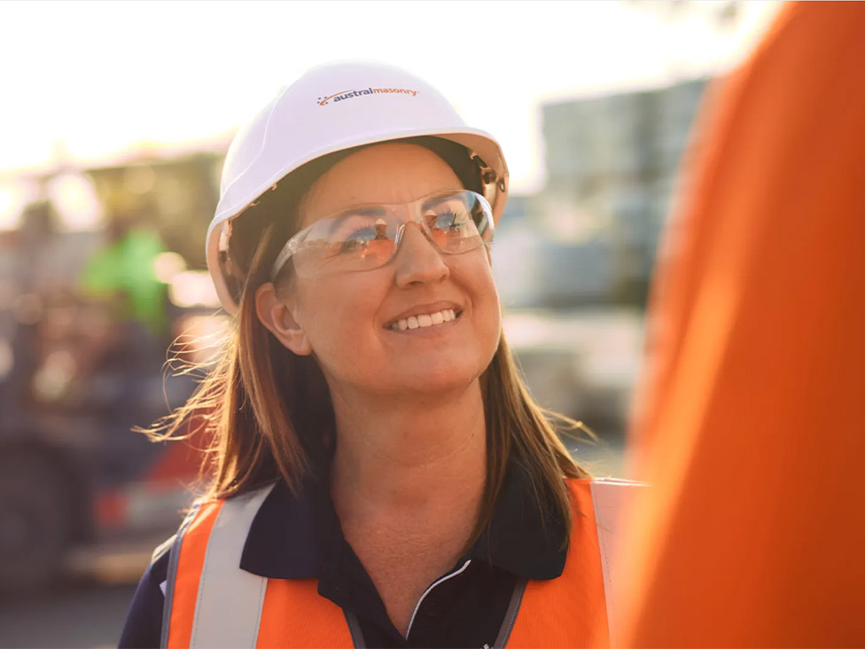 A woman wearing a hard hat, high-visibility vest, and safety glasses smiles outside on a sunny day.