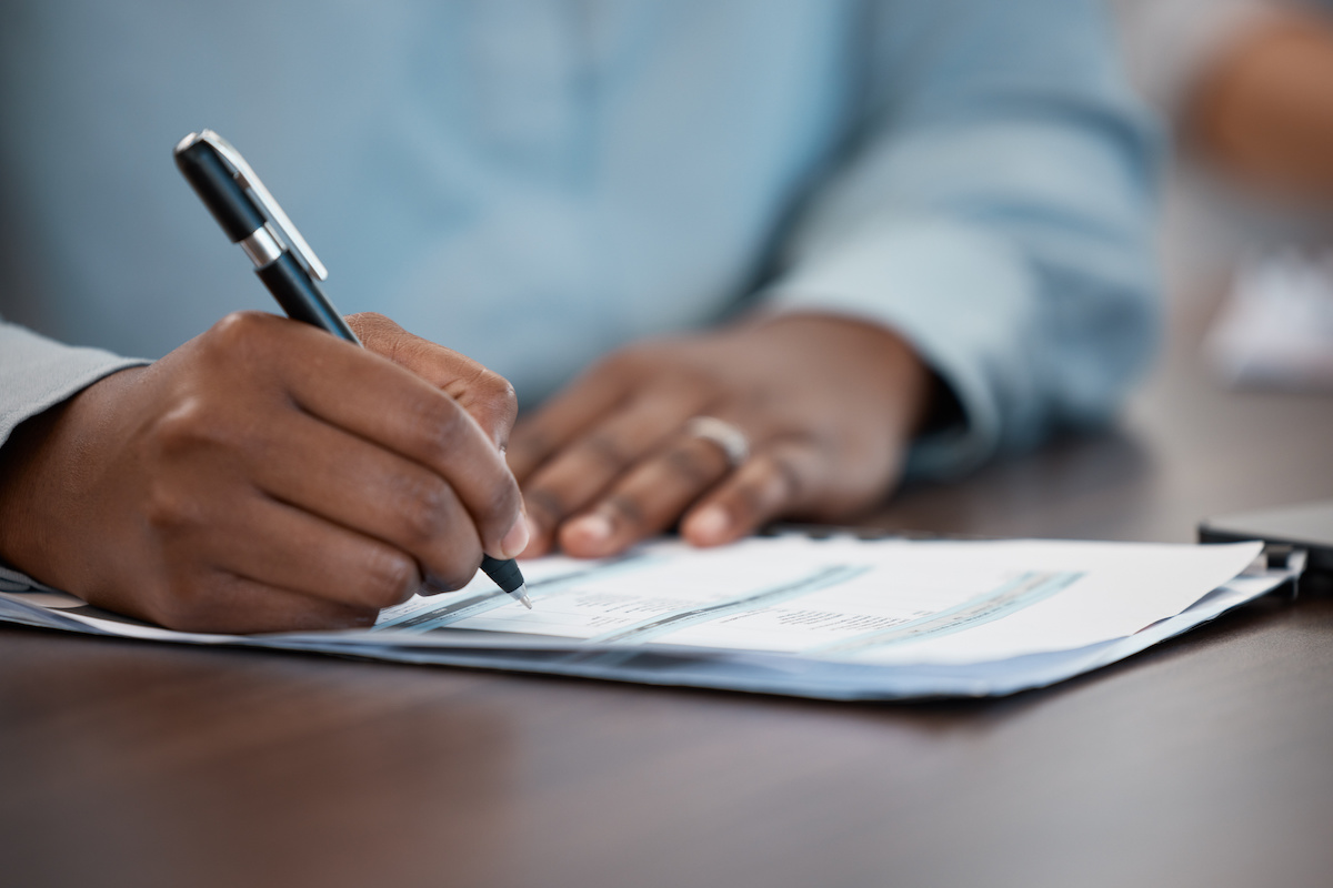 Hands, documents and contract with a black man signing paperwork at a table or desk in the office. Compliance, loan and insurance with a male putting his signature on a legal document for agreement.